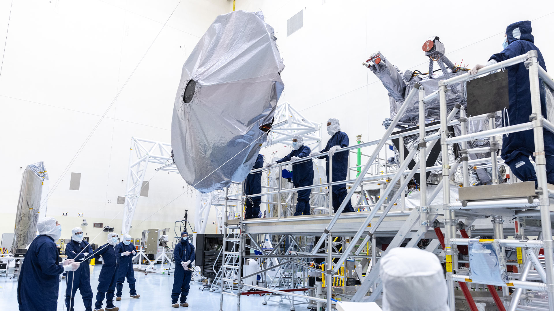 Workers in blue protective clothing and white head coverings hold cables to position Europa Clipper's high gain antenna for installation on the spacecraft. Some workers are on the ground, and some are up on a platform.