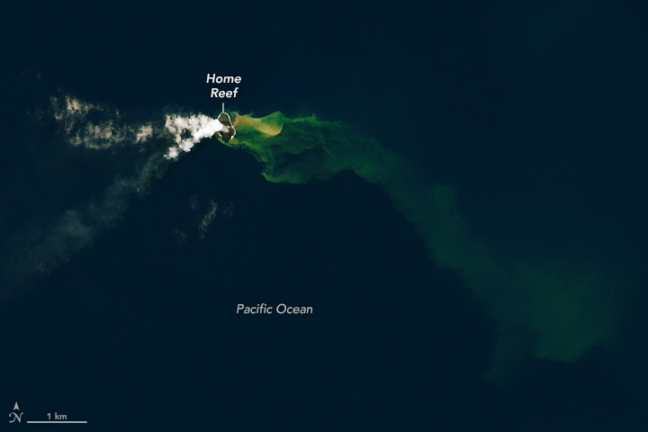 In a dark blue ocean there is a green cloudy tinge to the water emanating from the volcanic island peaking above the water in the upper left of center part of the image. Some small clouds emanate from the volcano and drift left in the atmosphere.