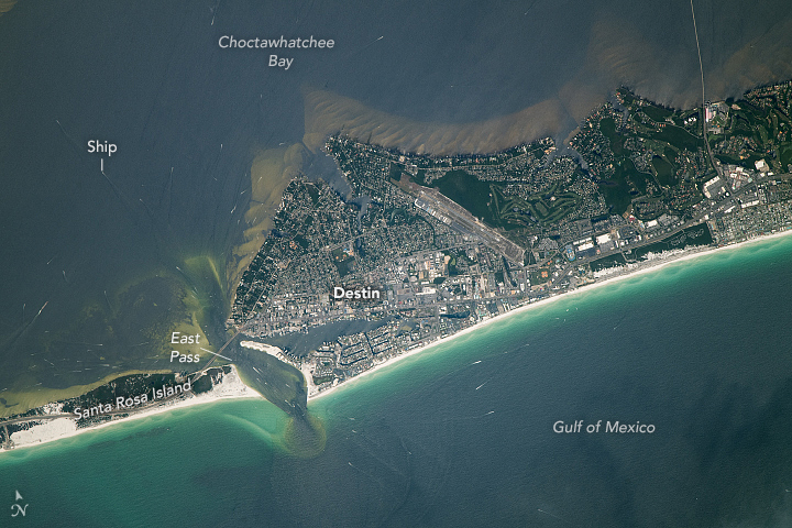 A barrier island splits this image horizontally, the more turquoise water of the Gulf of Mexico are seen at the bottom of the image and the more turbid water of Choctawhatchee Bay are seen above. The barrier island features an inlet where the land naturally narrows, but there is significant human settlement along the wider land to the right of the inlet.