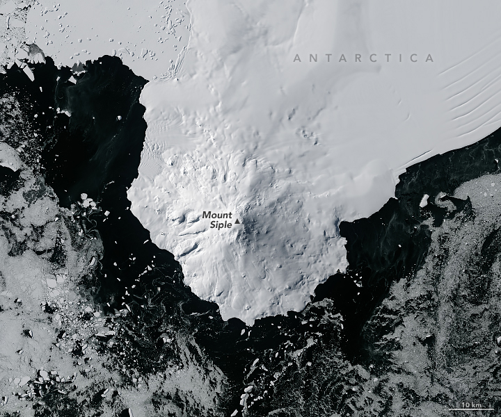 Dark, near black ocean surrounds the stark white ice sheet of a peninsula jutting down from the top of the image with clear topographic features compared to the ice sheet further up the image which is particularly smooth. There is also broken up icebergs floating in the water, however, they are mostly somewhat distant from the coastline.