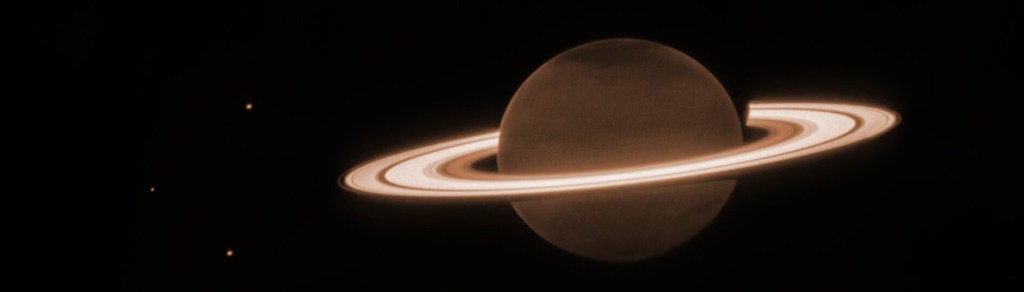 The background is mostly dark. At the center is a dark orange-brown circle, surrounded by several blazing bright, thick, horizontal whiteish rings. This is Saturn and its rings. There are three tiny dots in the image—one to the upper left of the planet, one to the direct left of the planet, and the lower left of the planet. These are three of Saturn’s moons: Dione, Enceladus, and Tethys, respectively. There is a slightly darker tint at the northern and southern poles of the planet. The rings surrounding Saturn are mostly broad, with a few singular narrow gaps between the broader rings. There is an innermost, thicker ring, and next to that is a brighter, wider ring. Traveling farther outward, there is a small dark gap before another thicker ring. In the thicker ring, there is a narrow faint band. There is then an outermost, faintest, thinnest ring.