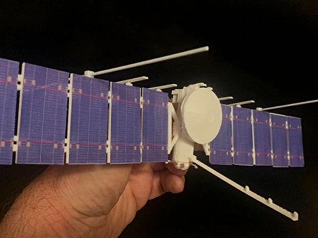 A hand holds a 3D printed version of Europa Clipper. The solar winds are bluish or purple. The rest of the spacecraft is pale yellow.