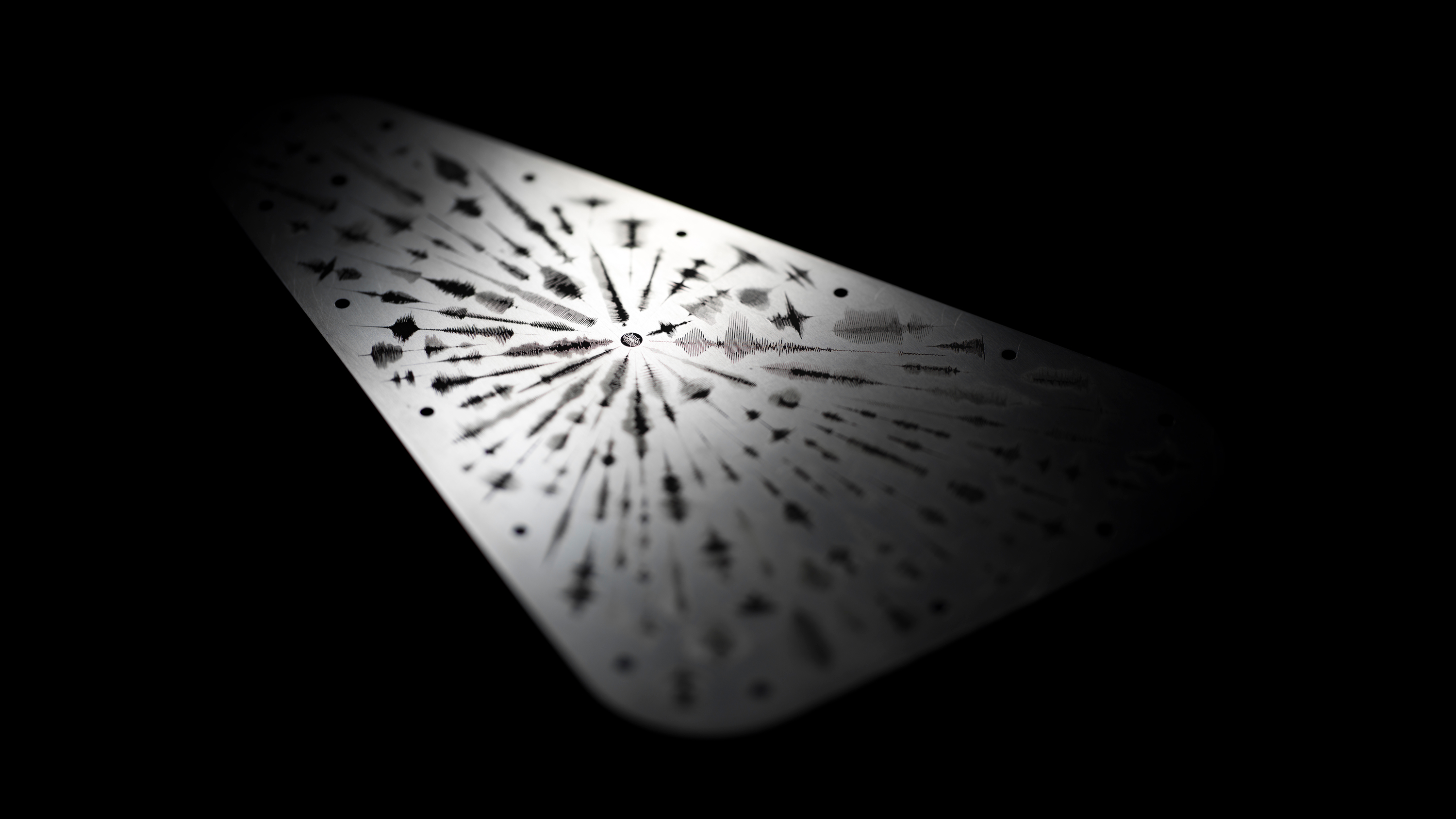 An image of the Europa Clipper vault plate, highlighted from the upper left corner casting shadows that emphasize engraved waveforms emanating from a central point symbolizing the word 'water' in multiple languages. The waveforms present as audio frequency waves. The vault plate is metal silver and the engravings are black, set against a black background.