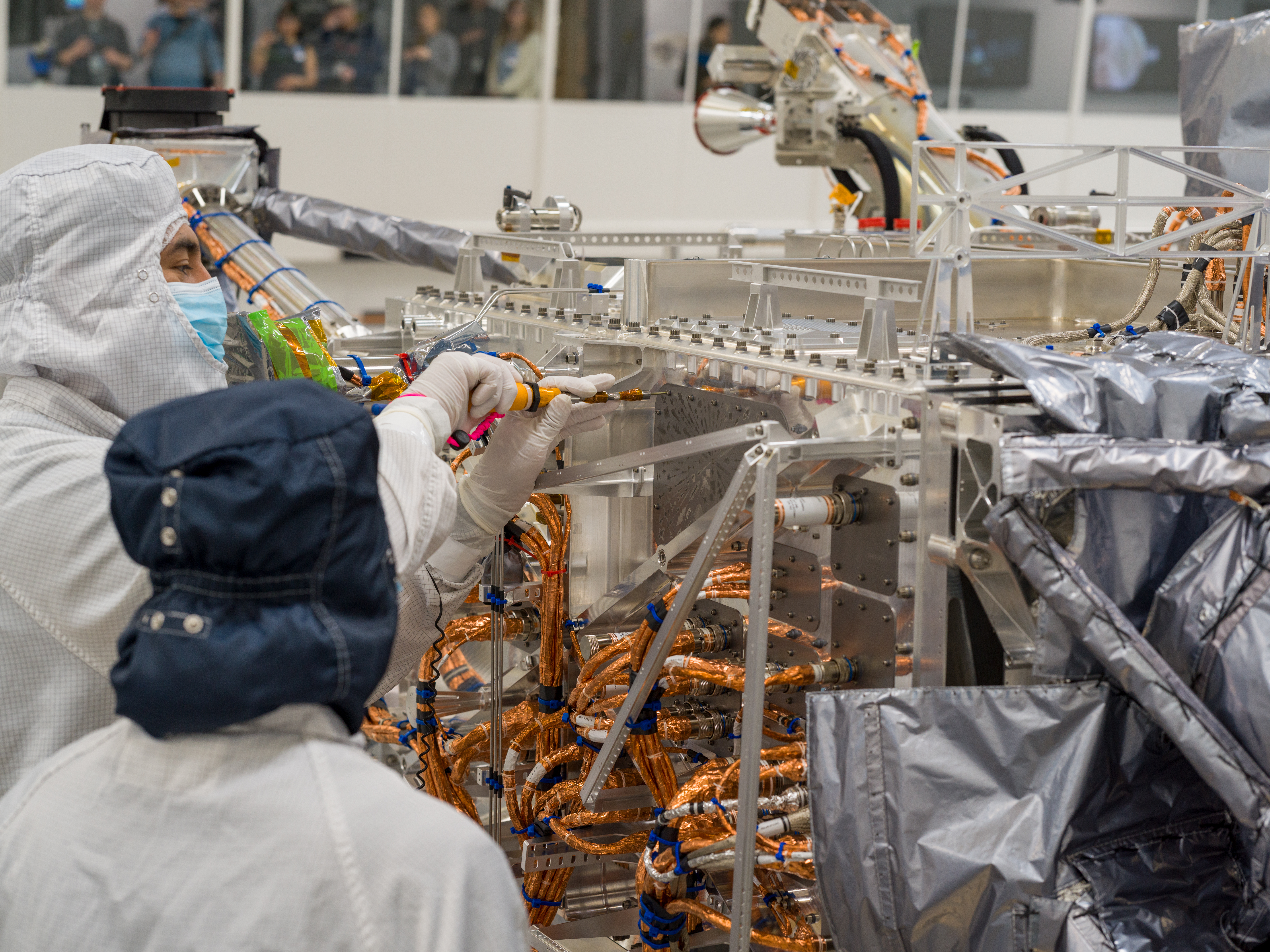A worker dressed in white protective clothing uses an orange-handled screwdriver to turn a screw attaching the triangular-shaped silver-colored vau...