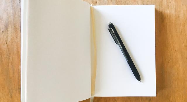 An open notebook with white paper sits on a wooden table. A pen is sitting on the right side of the notebook.