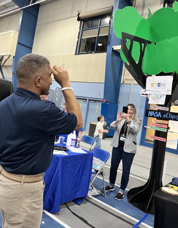 A man practices calculating the height of a tree using a clinometer to view the top of a 13-foot artificial tree while a NASA eClips volunteer takes his photo at the NASA eClips exhibit booth during CNU’s Community STEM Day.