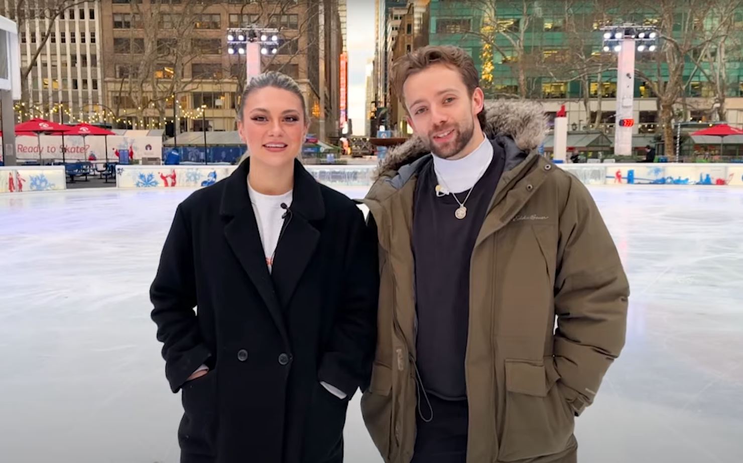 Two figure skaters wearing jackets stand on an ice rink.