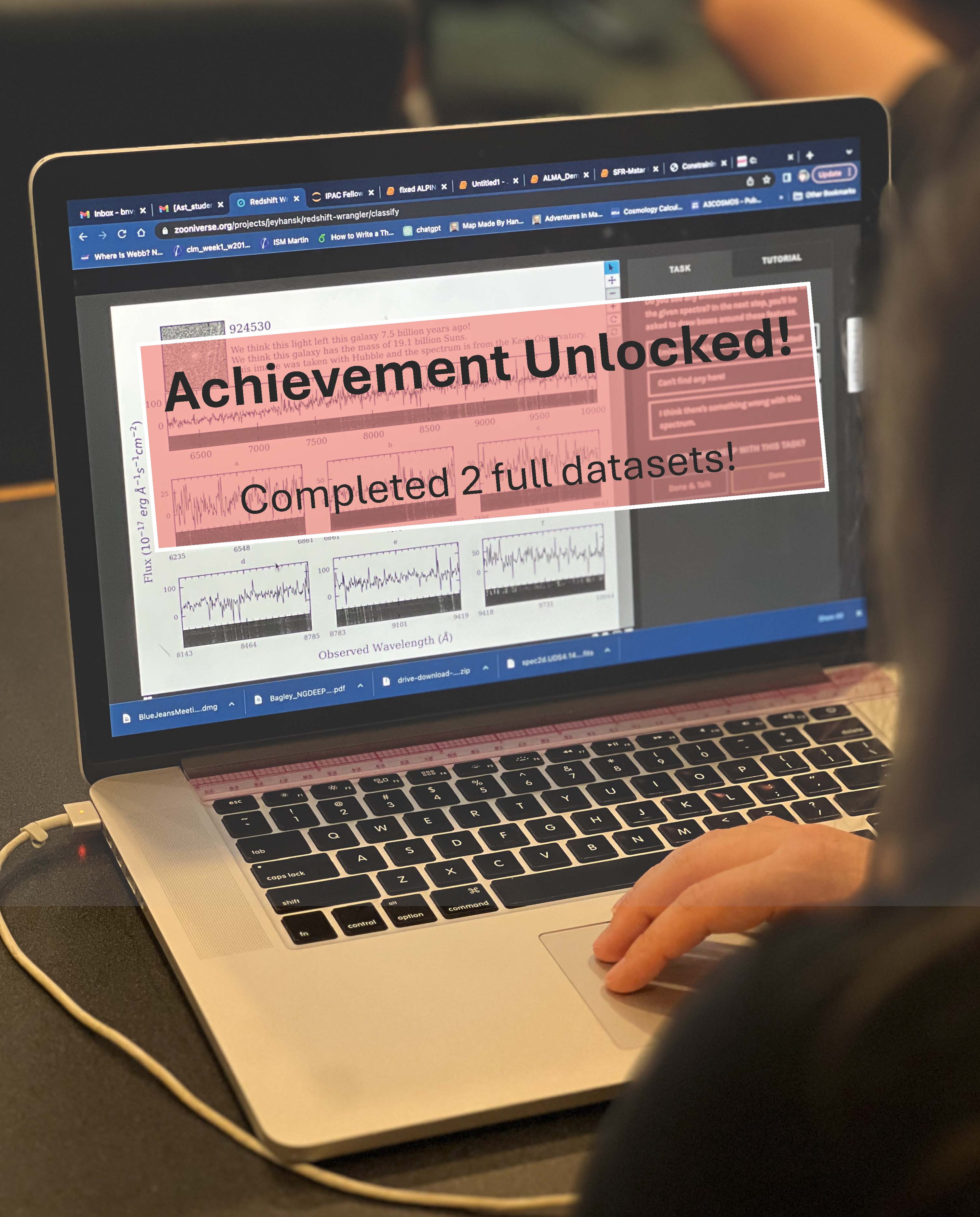 **Alt Text:** A person is working on a laptop with a screen displaying graphs and text. The screen has a large overlay with the text "Achievement Unlocked! Completed 2 full datasets!" The person's right hand is visible on the trackpad, and various browser tabs are open at the top of the screen, indicating an active and busy research session. The environment suggests a focus on data analysis and scientific work.