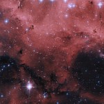 Glowing clouds of pink and red gas and dust fill the image, along with several stars – foreground stars shine with diffraction spikes, and more distant stars fill the background.